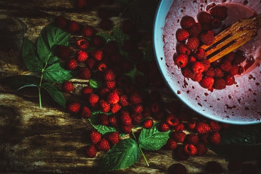 Red fresh raspberries on brown rustic wood background. Bowl with natural ripe organic berries with peduncles, green leaves and wooden fork, top view, flat lay with copy space.