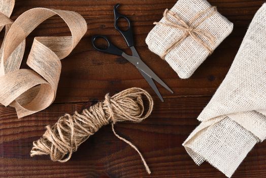 Top view of the tools for wrapping Christmas presents with burlap fabric and ribbon with scissors and twine on a dark wood surface.