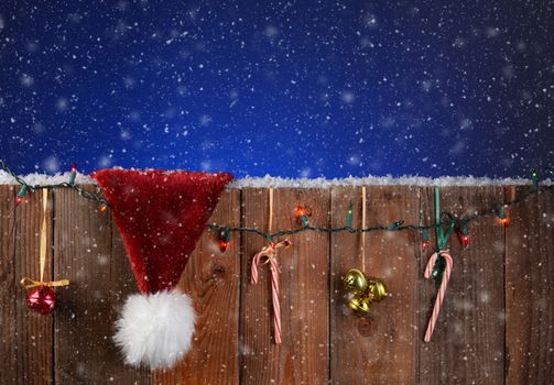 A rustic fence with christmas lights, bells, candy canes and a Santa Claus hat. Horizontal format with a light to dark blue night sky and snow effect. 