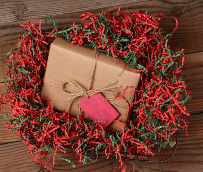 High angle view of a Christmas present wrapped with eco friendly craft paper and tied with twine. The package is resting on a field of red and green shredded crepe paper in a wire basket on rustic wood table.