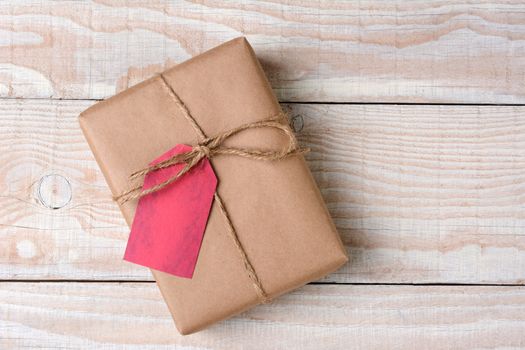 High angle shot of a plain brown paper wrapped Christmas present on a whitewashed wood table. The gift is tied with twine with a blank red gift tag.