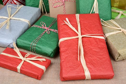 Closeup of a large group of wrapped Christmas presents on a burlap surface. A variety of presents wrapped with tissue paper and plain paper tied with string and raffia, Horizontal format.
