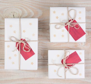 High angle image of Christmas presents wrapped in white paper and tied with white string. Red gift tags and stars adorn the packages on a white wood table. Square format.