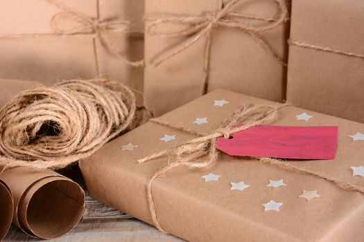 Closeup of a group of Christmas packages wrapped with plain brown paper and twine. Horizontal format on a wood table. A red gift tag and paper stars adorn one parcel.