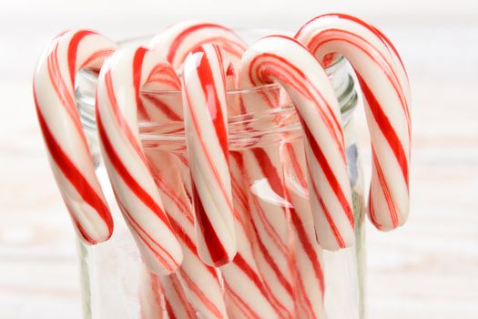 Closeup of a bunch of holiday candy canes hooked on the rim of a glass jar. Shallow depth of field on a whitewashed wood background. Horizontal format.