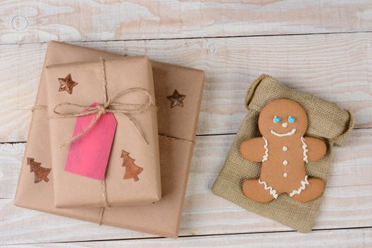 High angle shot of two Christmas presents and a gingerbread man on a whitewashed wood table. The cookie is on a burlap bag. 