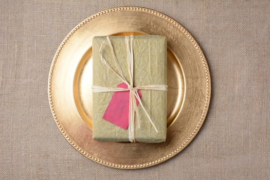 High angle shot of a tissue paper wrapped Christmas present on a gold charger on a burlap surface. The gift has a blank gift tag and is tied with raffia. Horizontal format.