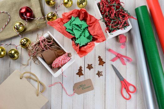 Overhead view of Christmas wrapping supplies on a rustic wood table. Scissors, ribbon, bells, tags, paper rolls, gift bag, string, ornaments and crepe paper are displayed.