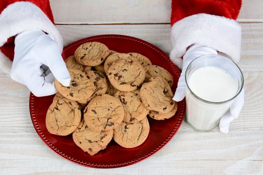 Closeup of Santa Claus with a plate full of chocolate chip cookies and a glass of milk. Only Santa's hands are visible. Horizontal format from a high angle.