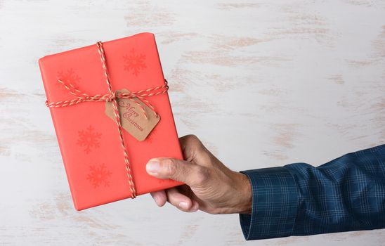 Closeup of a man in hand holding a Christmas present wrapped with red paper over a rustic white wood background. Horizontal format with copy space.