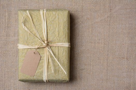 Christmas present wrapped with gold tissue paper, tied with raffia and a blank gift tag. On burlap with copy space.