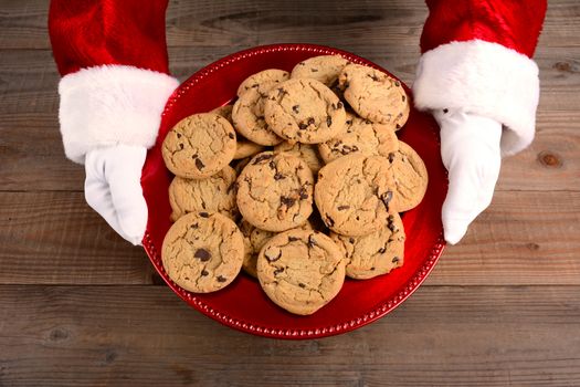 Closeup high angle view of Santa Claus serving cookies. Only santa's hands are shown as he holds a red tray filled with chocolate chip cookies over a rustic wood table.