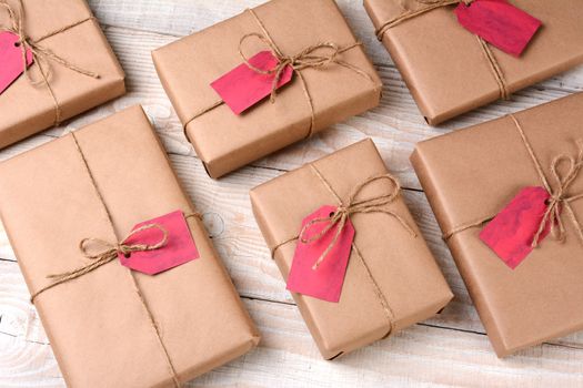 A group of plain brown paper wrapped Christmas presents on a white wood table. The gifts are tied with twine and have blank red gift tags. High angle shot.