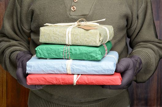 A man wearing sweater and gloves holding a stack of wrapped Christmas presents in front of his torso. Man is unrecognizable. Horizontal format.