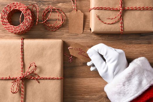 High angle view of Santa Claus hand holding a gift tag with Merry Christmas. Closeup with wrapped presents and string.