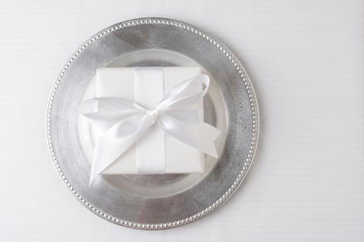 High angle shot of a gift wrapped in white paper and tied with a white ribbon. The present is in the middle of a silver charger on a white tablecloth. Horizontal format with copy space.