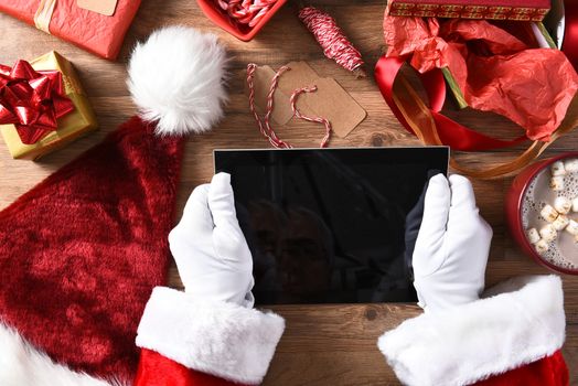 Santa Claus holding a generic tablet computer over a table filled with presents, wrapping paper, tags and a mug of cocoa. Horizontal format showing hands only.