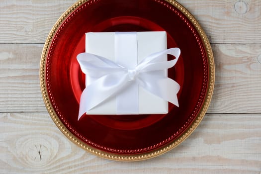 High angle shot of a present wrapped in white paper and tied with white ribbon and bow. The present is centered on a red charger on top of a gold charger. Horizontal format on white wood table.