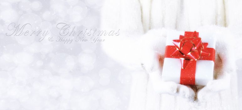 Closeup of a woman with a present in her hands with Merry Christmas and Happy New Year on a silver bokeh background and snow effect. Banner size with copy space.