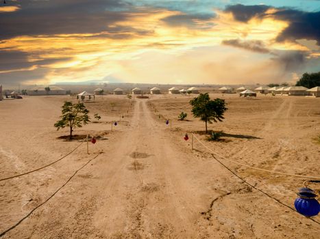 Empty dirt road with barren desert on both sides and blue sky above showing thar desert of rajasthan india. Shows the tents of a desert camp in the distance with lanterns hanging on the side