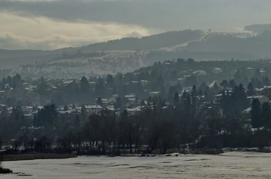Winter scene with frozen lake, snowy mountain, glade, forest and residential district of bulgarian village Pancharevo, Sofia, Bulgaria