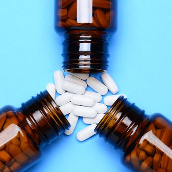 A closeup of three prescription medicine bottles laying on their sides with pills spilling onto a blue surface. Overhead shot with the bottles as the main focus.