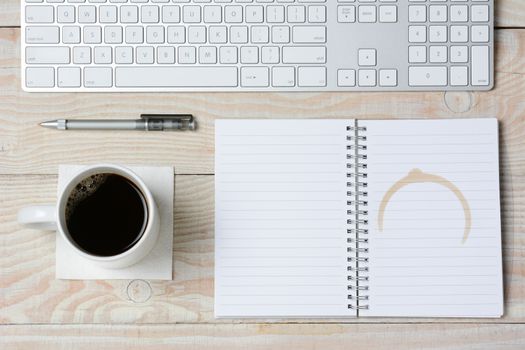 High angle shot of a white rustic desk with a modern keyboard, coffee cup and notebook with a coffee stain. Horizontal format.