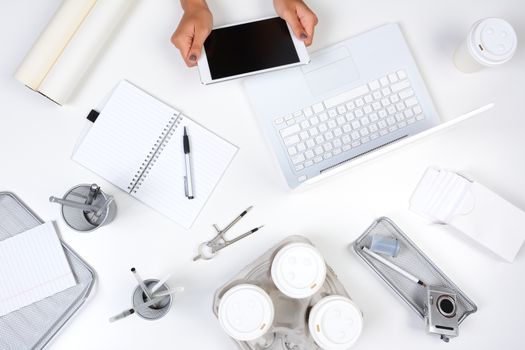 High angle shot of a white desk with primarily white and silver office objects, with a woman holding a tablet computer showing her hands only. Tablet has a blank screen.