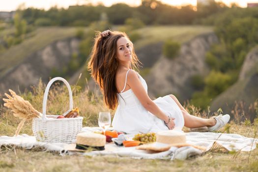 Gorgeous young brunette girl in a white sundress having a picnic in a picturesque place. Romantic picnic.