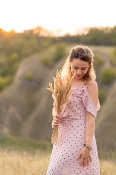 Beautiful tender girl in a white sundress walks at sunset in a field with a spikelet bouquet