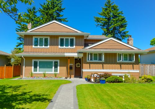 Average residential house in perfect neighbourhood. Family house with big green lawn and trees in front on blue sky background