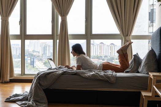 A beautiful young brunette girl is working on the laptop while lying on a bed by a panoramic window with a beautiful view from a high floor. Stylish modern interior. A cozy workplace.