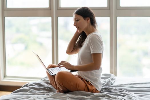 A beautiful young brunette girl is working on the laptop while sitting on a bed by a panoramic window with a beautiful view from a high floor. Stylish modern interior. A cozy workplace.