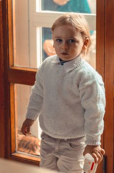 handsome blond little boy stands near the window with yogurt in his hand