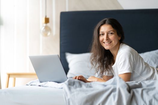 Beautiful young brunette girl with a laptop lying on the bed. Stylish modern interior. A cozy workplace. Shopping on the Internet