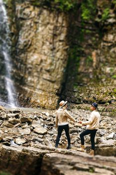 Young and beautiful couple at the mountain waterfall - Happy tourists visiting mountains. Lovestory. Tourists in hats. Military fashion