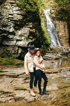 Young and beautiful couple at the mountain waterfall - Happy tourists visiting mountains. Lovestory. Tourists in hats. Military fashion