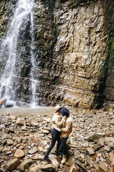 Young and beautiful couple at the mountain waterfall - Happy tourists visiting mountains. Lovestory. Tourists in hats. Military fashion