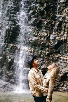 Young and beautiful couple at the mountain waterfall - Happy tourists visiting mountains. Lovestory. Tourists in hats. Military fashion