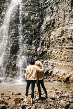 Young and beautiful couple at the mountain waterfall - Happy tourists visiting mountains. Lovestory. Tourists in hats. Military fashion