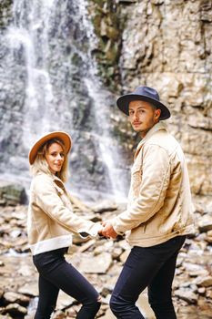 Young and beautiful couple at the mountain waterfall - Happy tourists visiting mountains. Lovestory. Tourists in hats. Military fashion