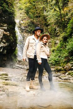 Young and beautiful couple at the mountain waterfall - Happy tourists visiting mountains. Lovestory. Tourists in hats. Military fashion