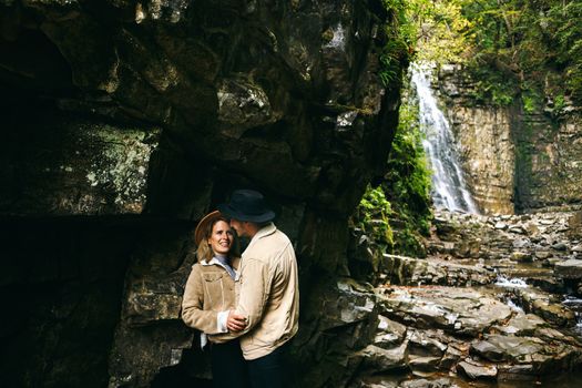 Young and beautiful couple at the mountain waterfall - Happy tourists visiting mountains. Lovestory. Tourists in hats. Military fashion