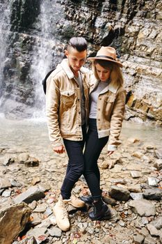 Young and beautiful couple at the mountain waterfall - Happy tourists visiting mountains. Lovestory. Tourists in hats. Military fashion