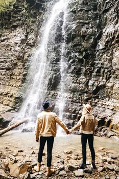 Young and beautiful couple at the mountain waterfall - Happy tourists visiting mountains. Lovestory. Tourists in hats. Military fashion