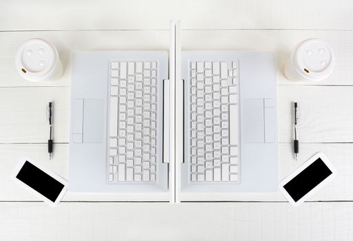 High angle shot of two laptop work stations back-to-back with almost identical layouts. Horizontal format on a white wood table.
