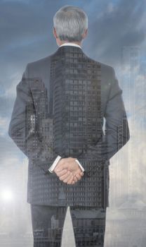 Double exposure of a businessman with his hands clasped behind his back with city background.