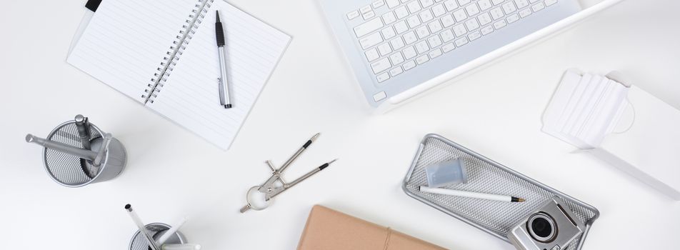 High angle shot of a white desk with primarily white and silver office objects. Items include, laptop, computer, pad, pens, camera, with a brown wrapped package at the bottom of the frame. Banner Format.