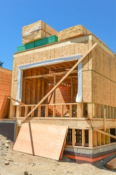 A single family home under construction. The house has been framed and covered in plywood. Stacks of board timber in front and stack of 2x4 boards on the top.