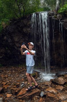 Stunning view of a boy tourist taking selfie photos with a smartphone at the waterfalls background in the Altai mountains.